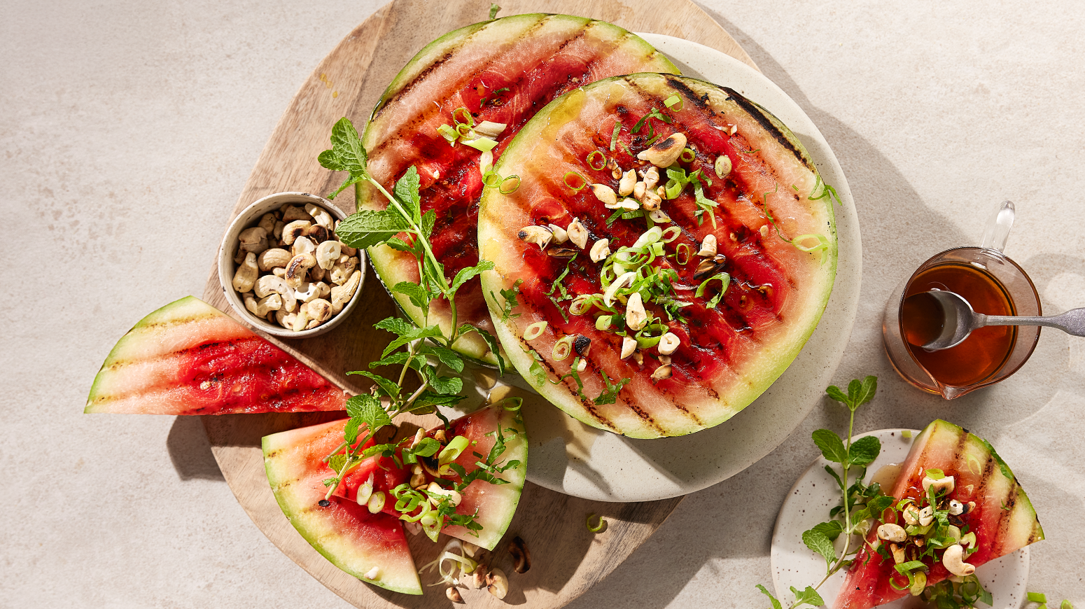 Grillet vannmelon med vårløk og cashewnøtter, servert på hvit tallerken på lys trefjøl. Dressing i en glasskopp ved siden av.