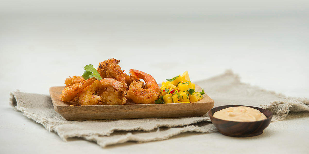 Kokosreker; stekte scampi i brødsmuler og revet kokos, servert med mangosalat på trefat, på linserviett. Liten treskål med chilimajones ved siden av.