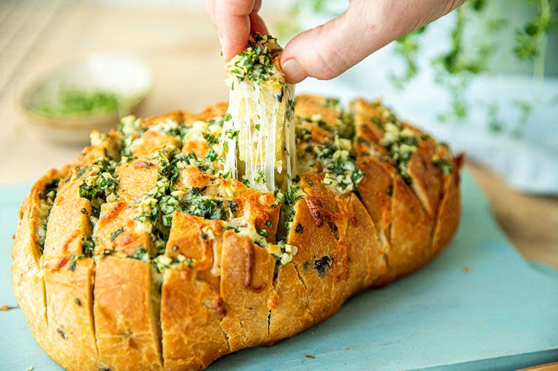 Pull apart -hvitløksbrød med smeltet ost og urter, laget av et helt landbrød. Hånd som drar ut en rute med brød, med smeltet ost som strekker seg fra det hele brødet. Lys blå bakgrunn.