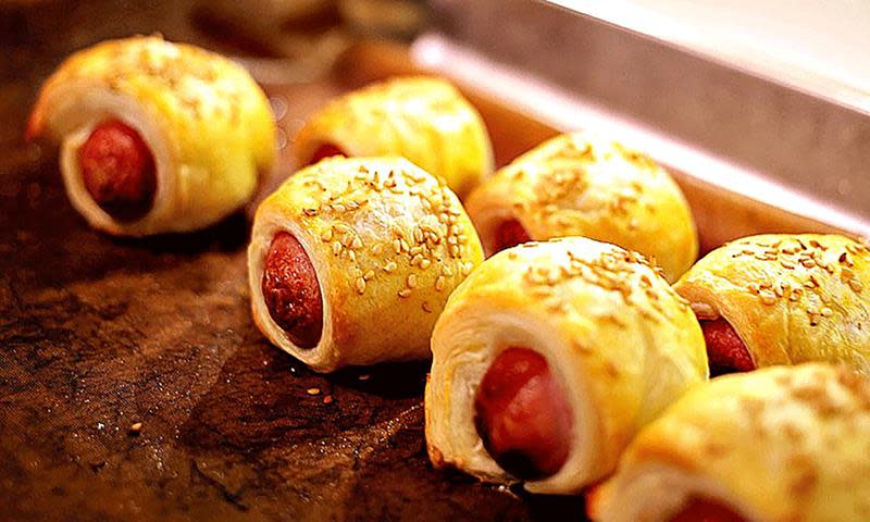 Innbakte pølser med butterdeig og sesamfrø, på rad og rekke på mørkt underlag.