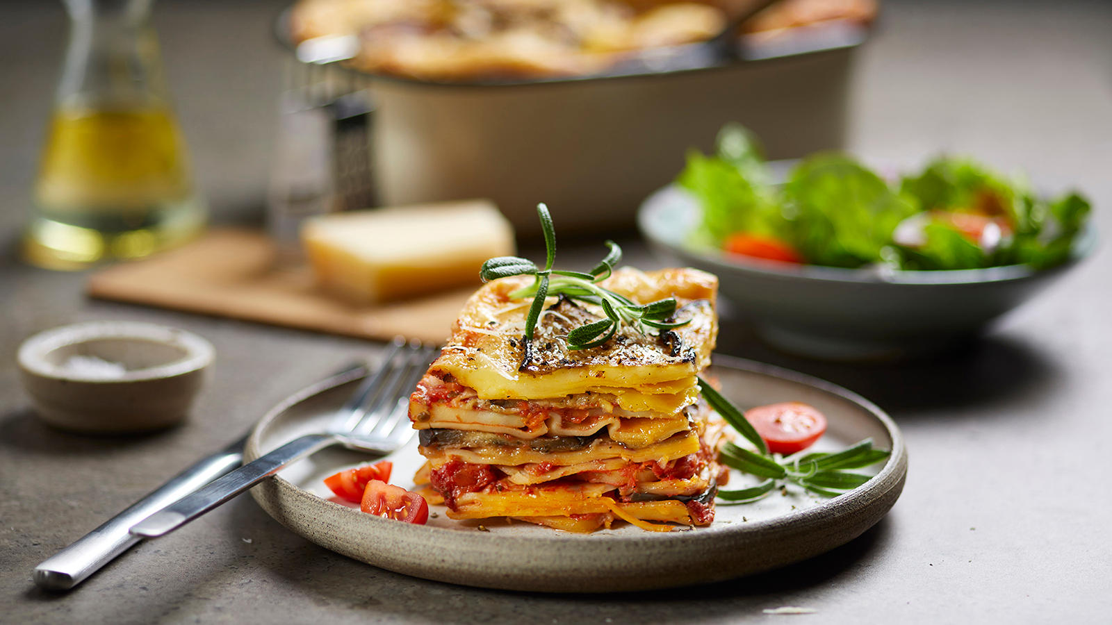 Saftig vegetarlasagne med aubergine og rosmarin delt i stykker og servert på lysegrå asjett. Skål med salat, trefjøl med parmesan og ildfast form med lasagne i bakgrunnen.