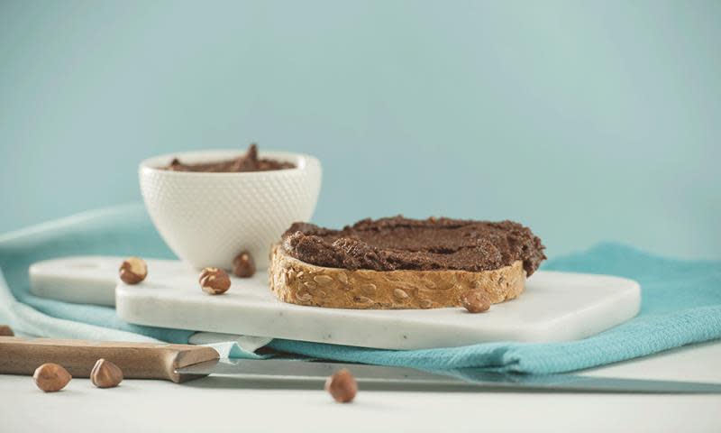 Sunt sjokoladepålegg med hasselnøtter smurt utover en brødskive, servert på liten hvit marmorfjøl. Hasselnøtter rundt brødskiven, og en hvit skål med sjokoladepålegg i bakgrunnen.