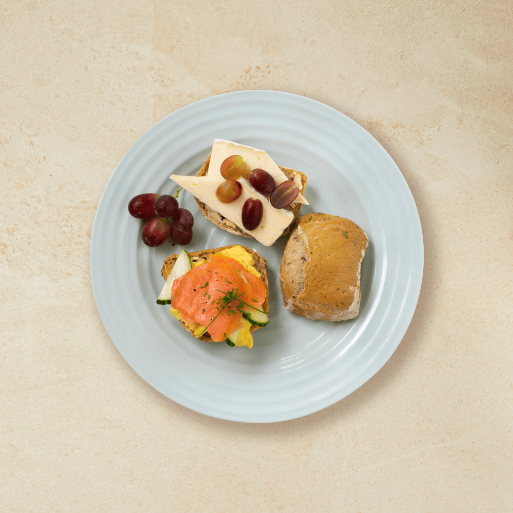 Halvgrove eltefrie rundstykker servert med eggerøre, røkelaks, brie og druer på en lys tallerken.