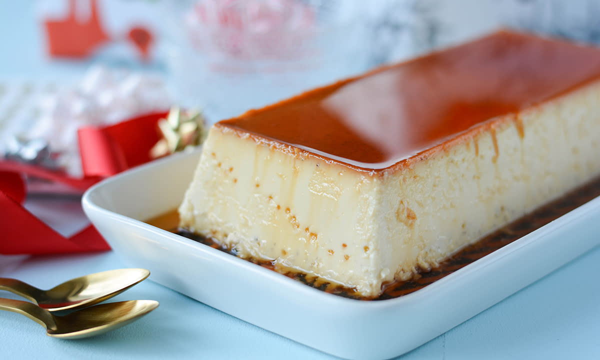 Hjemmelaget karamellpudding i hvit avlang form med røde bånd og gullskjeer ved siden av.