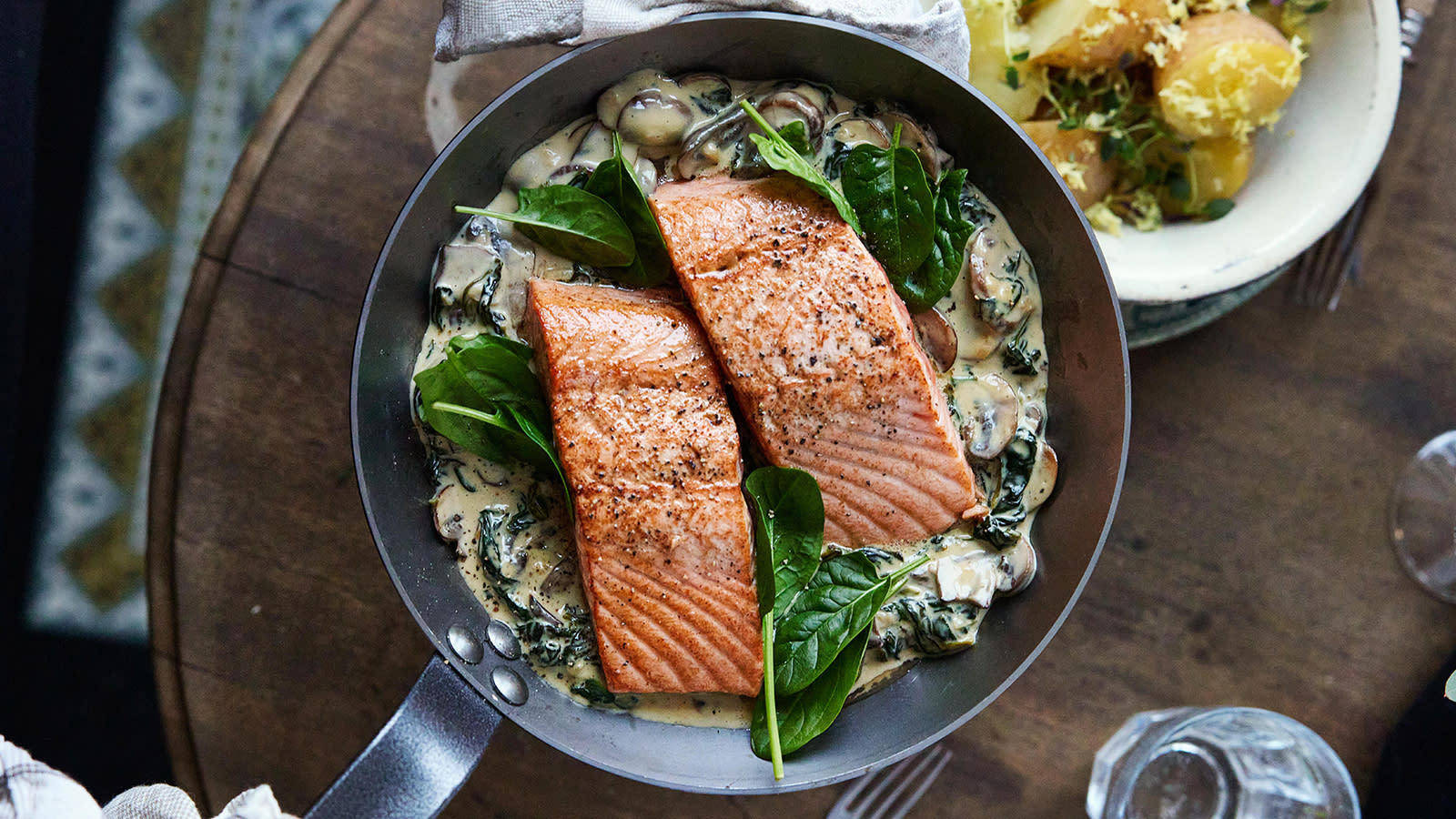 Pannestekt laks med lun potetsalat og kremet sopp- og spinatsaus servert i metallpanne med lun potetsalat ved siden av.