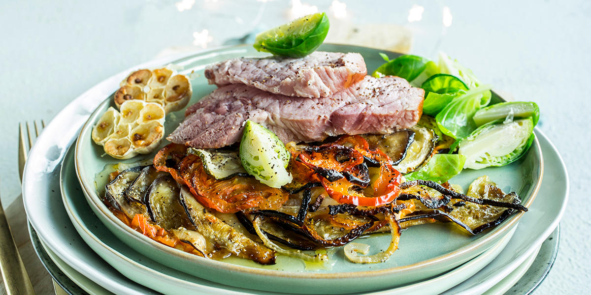 Lys grønn tallerken med skiver av badsturøkt lammelår, ovnsbakte grønnsaker, rosenkål og halve ovnsbakte hvitløk. Diskret julelys i bakgrunnen.