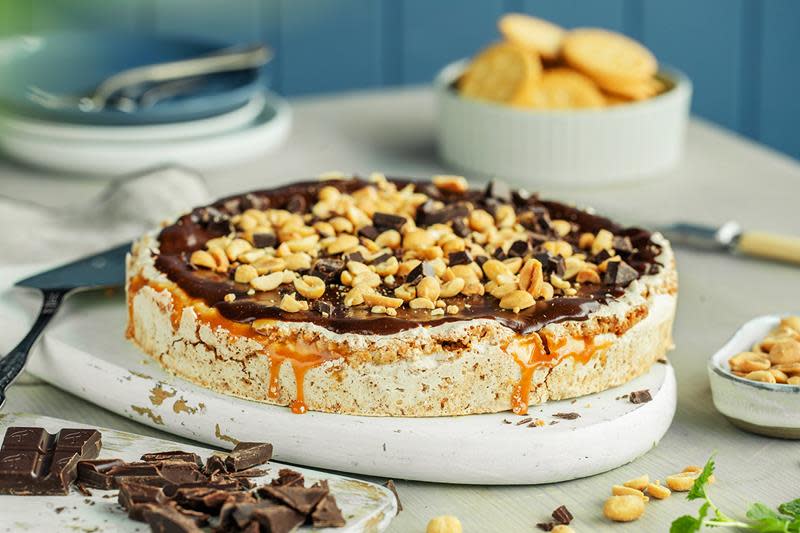 Snickerskake med peanøtter og karamell servert med sjokoladekrem på lys trefjøl. Skål med peanøtter ved siden av, tallerkener i bakgrunnen og hakket sjokolade i forgrunnen.
