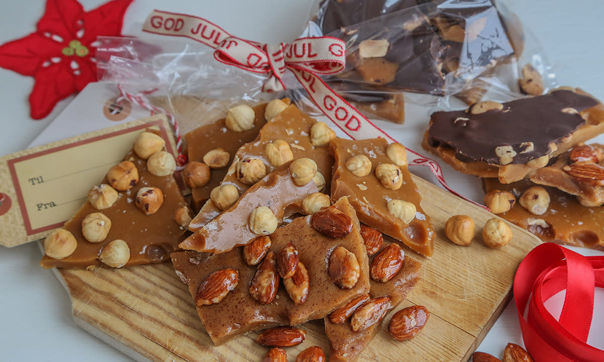 Kramellknekk med forskjellig nøttetopping og sjokolade, servert på trefjøl. Juleinnpakket karamellknekk i cellofanposer i bakgrunnen.
