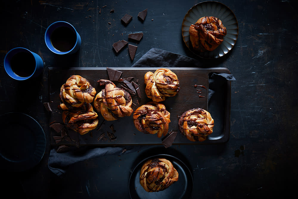 Kanelsnurrer med sjokolade på et mørkt fat, servert med sjokoladesaus ringlet over, og to kopper kaffe på siden.