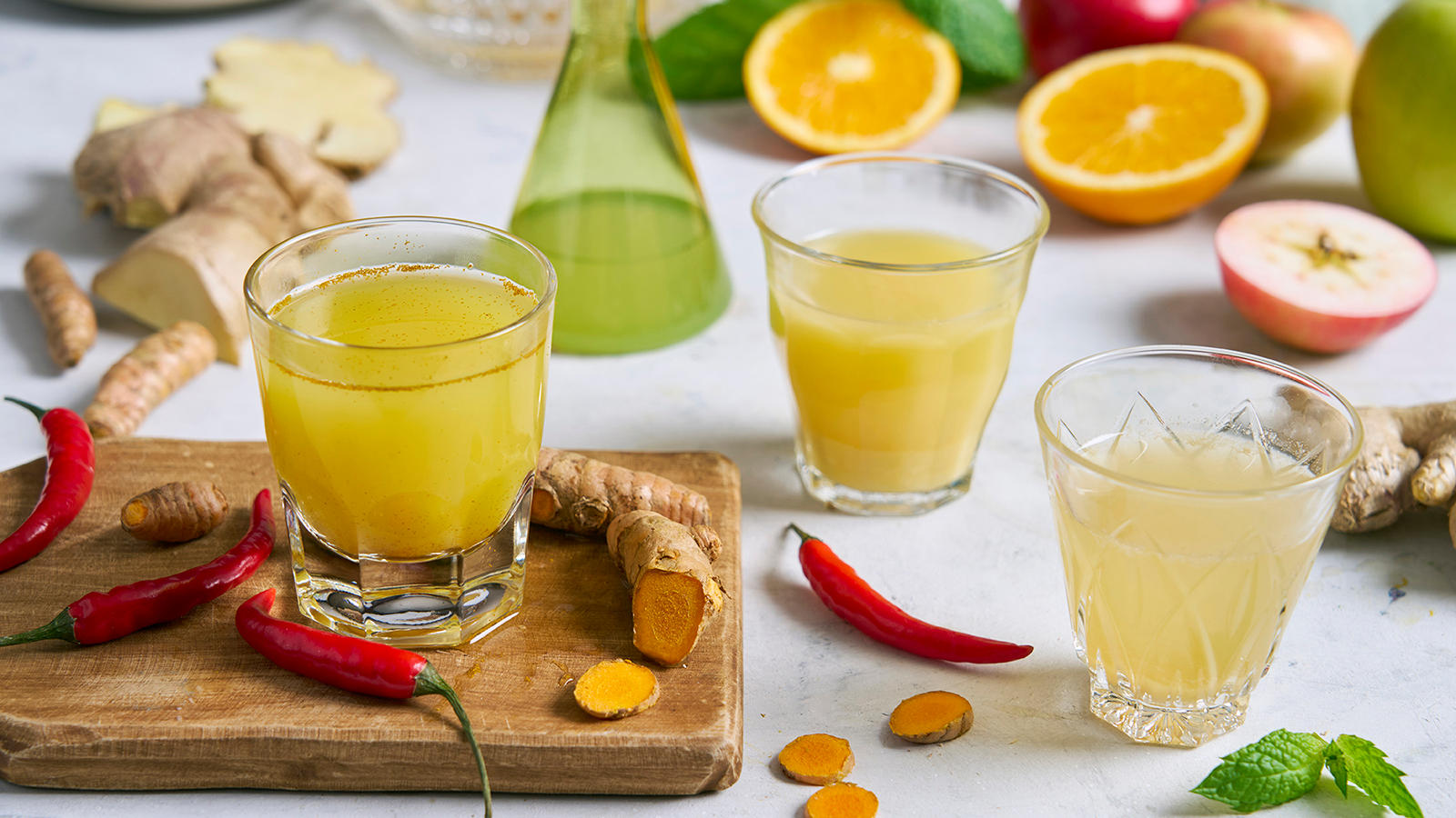Hjemmelaget ingefærshot med forskjellige smaker servert i tre forskjellige glass. Friske biter av ingefær, appelsin, eple, gurkemeie og hele røde chili ligger rundt glassene.
