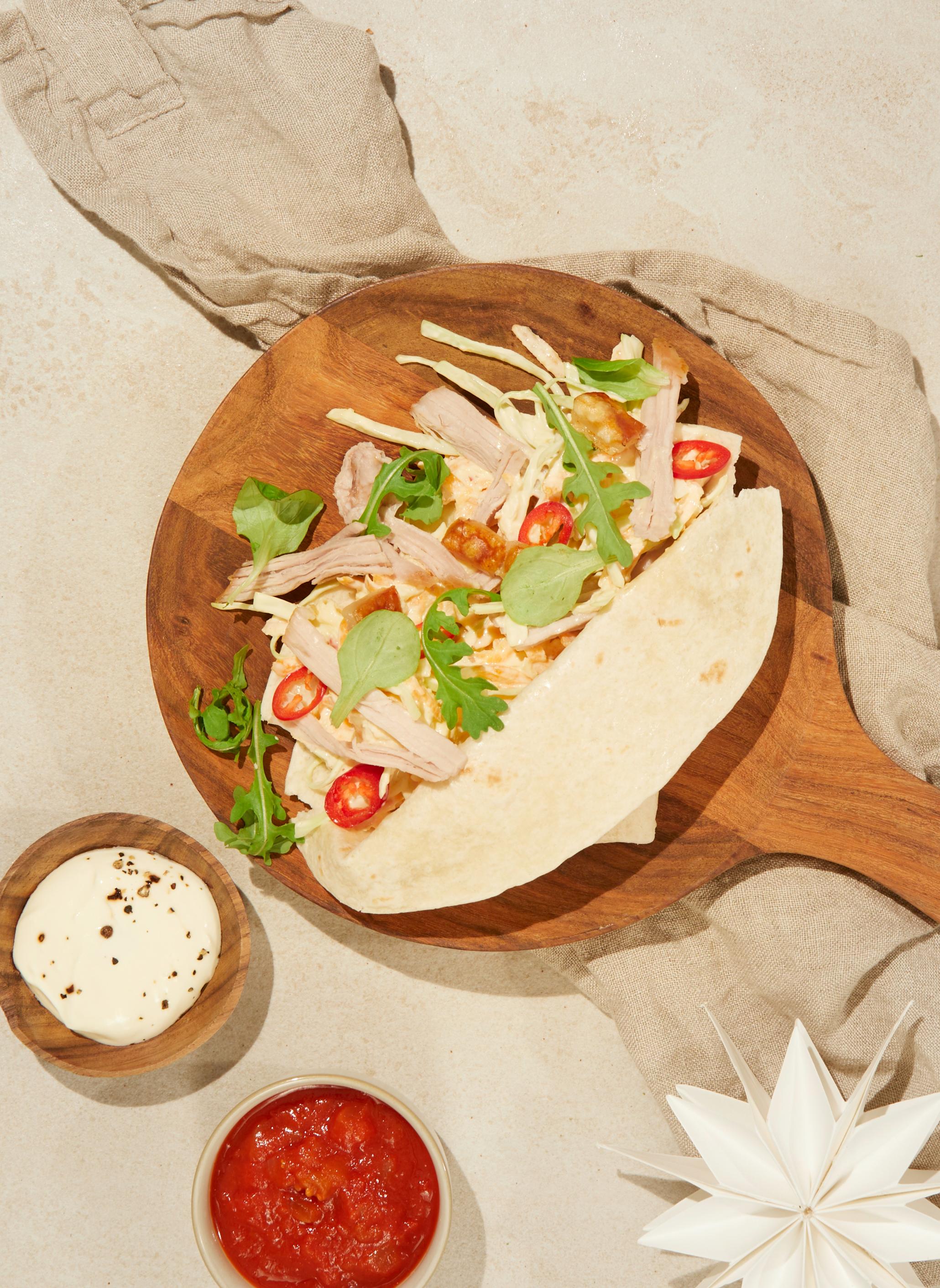 Pulled pork taco med ribberester og hjemmelaget coleslaw servert i tortillalefse på mørk trefjøl, med beige linhåndkle under. På siden er det to små skåler med rømme og salsa.