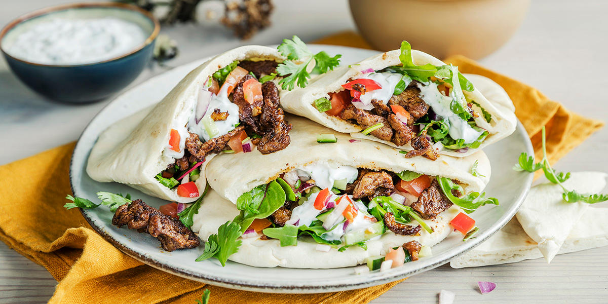Fat med tre pitabrød fylt med lammeskav, tomater, urter og yoghurtdressing, servert på lys tallerken på gul linserviett. Liten skål med yoghurtdressing i bakgrunnen.