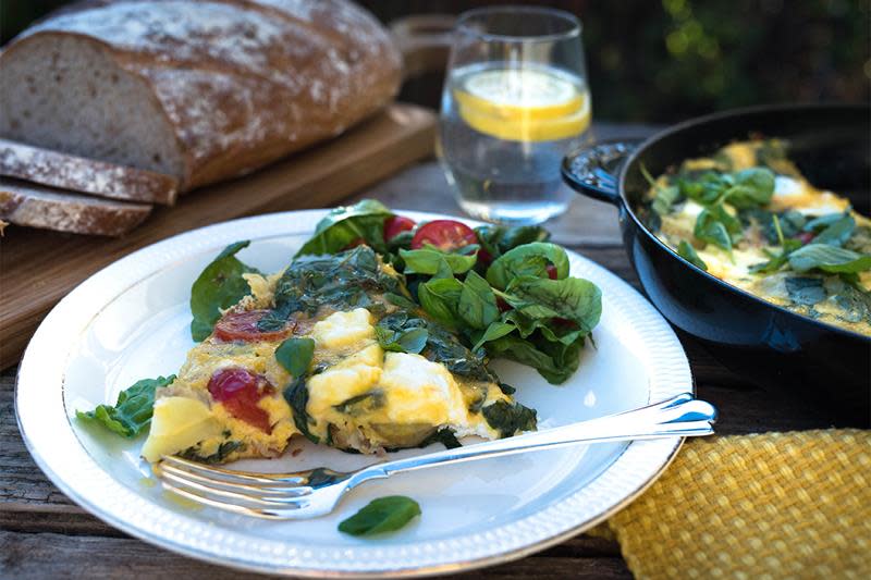 Et stykke bondeomelett med tomater, bacon, chèvre og fersk basilikum servert på hvit tallerken, med fersk salat ved siden av. Brød i bakgrunnen, vannglass med sitronskiver og jernpanne med bondeomelett på rustikk trebord.