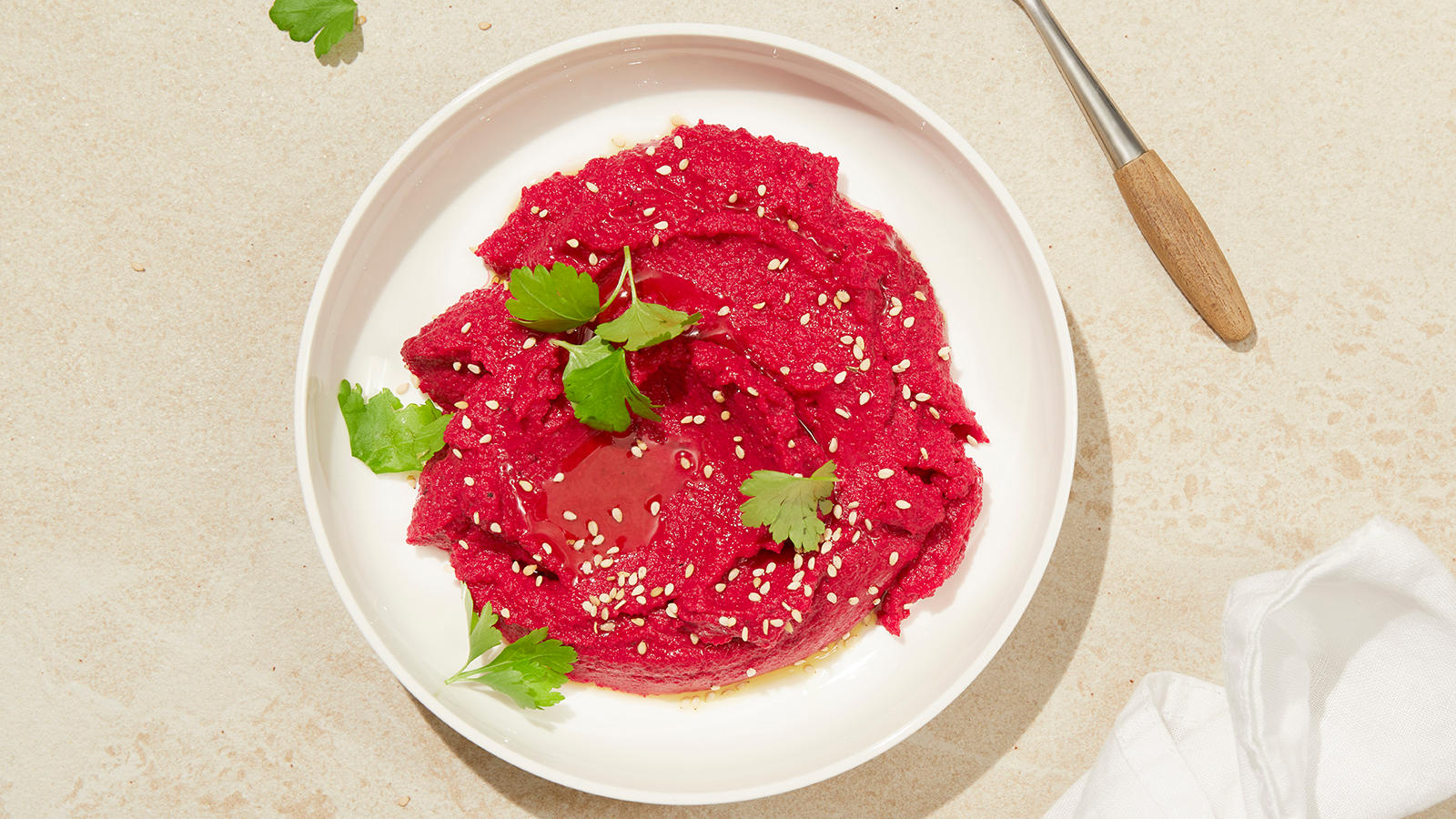 Hummus med rødbeter, servert i hvit skål med sesamfrø, olivenolje og bladpersille på toppen.