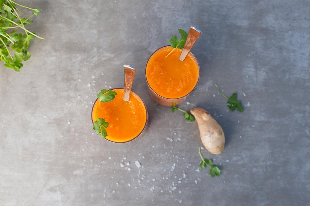 Gulrotsmoothie med ingefær og mango, servert i to glass på mørk grå bakgrunn. Fersk ingefær og friske urter strødd rundt.