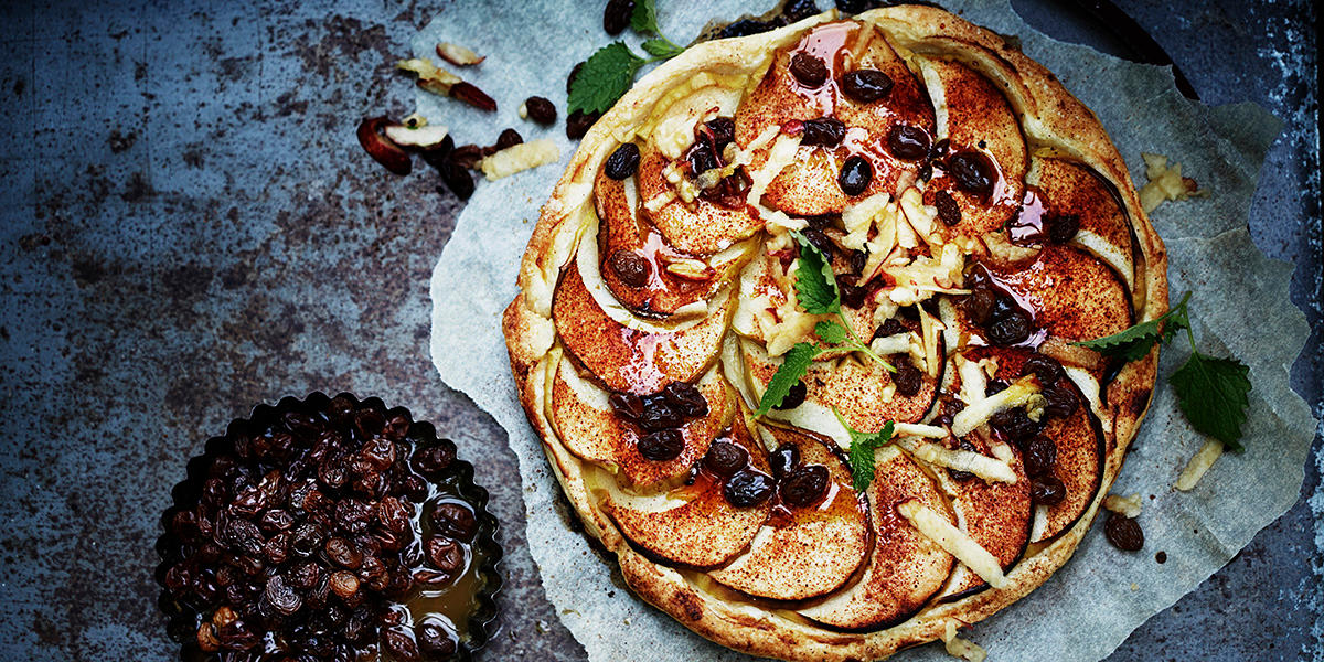 Enkel eplepai med butterdeig og skivede epler, toppet med rosiner, grønne urter og kanel. Servért på bakepapir på mørk bakgrunn. Liten skål med eplekokte rosiner ved siden av.