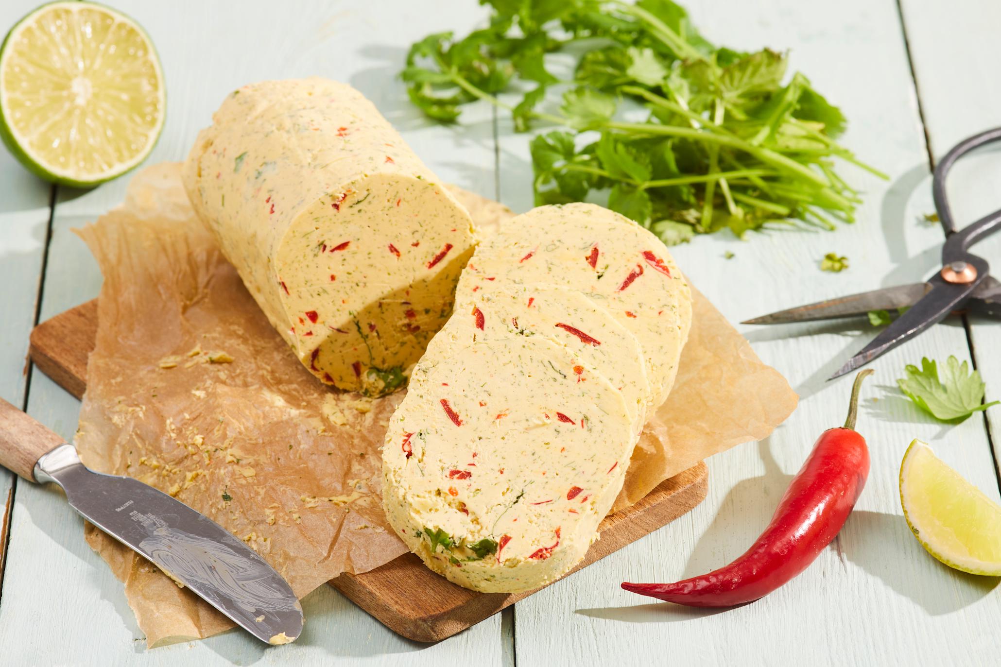 Hjemmelaget hvitløkssmør med chili og lime servert i skiver på liten trefjøl med bakepapir. Friske urter, urtesaks, limebåt og chili i bakgrunnen og på siden.