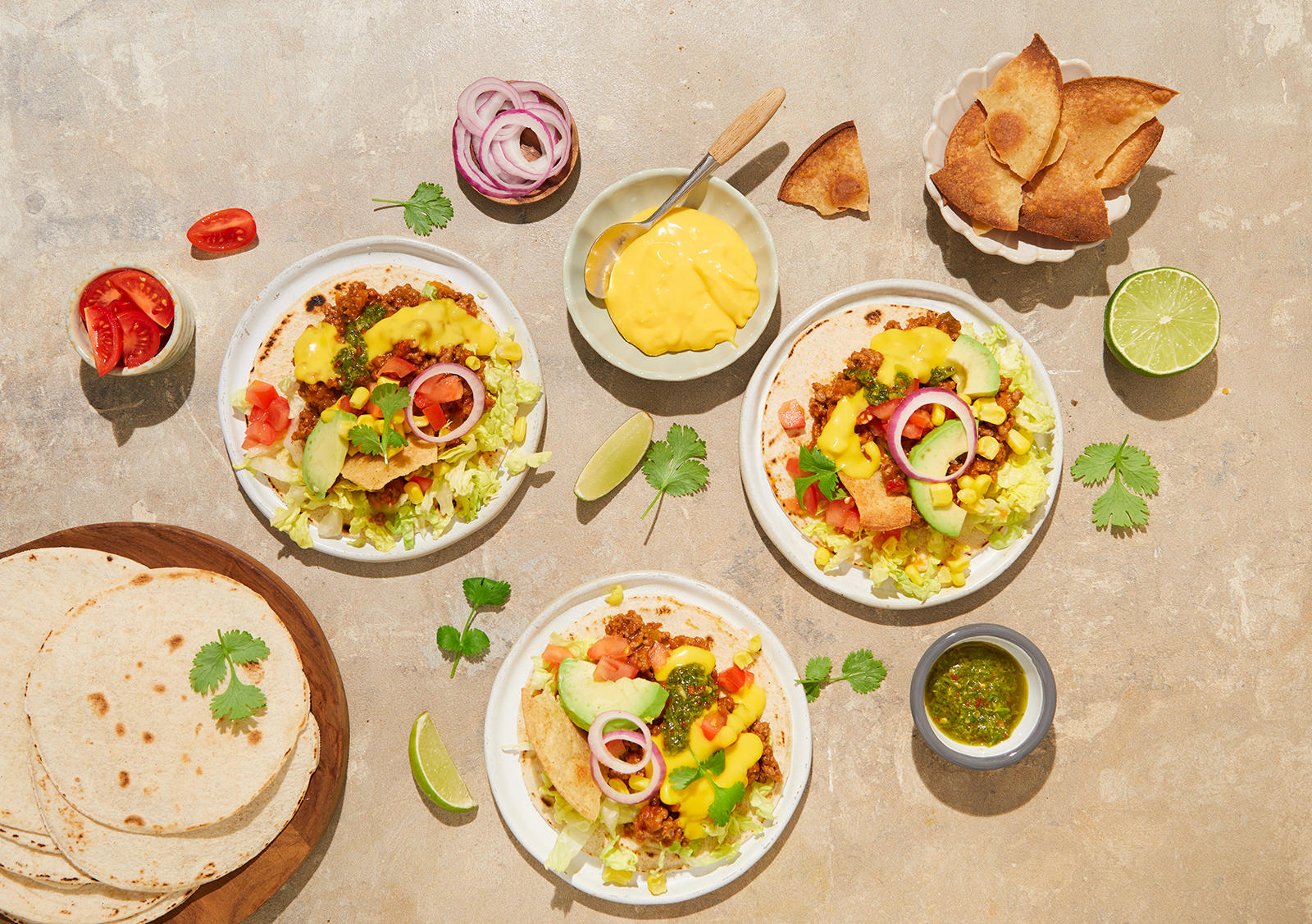 Klassisk taco med kjottdeig, chimichurri og kavli cheddar dip servert på små hvite tallerkner med limebåter, frisk koriander, tomater og rødløk rundt. Liten skål med krydrede tortillachips på siden.
