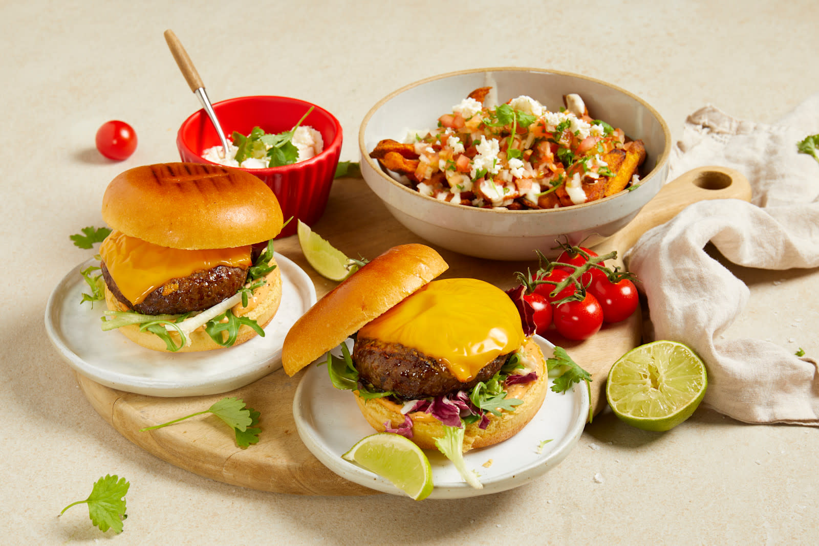 Big Juicy burger med cheddar og loaded søtpotetfries servert på to små hvite asjetter med limebåter og cherrytomater rundt. Skål med loaded søtpotetfries med tomatsalsa, hvitløksdressing og fetaost i bakgrunnen.