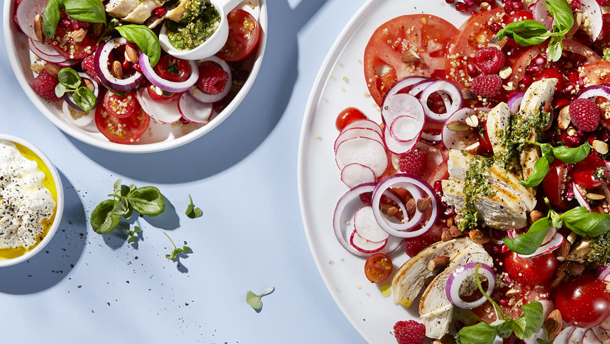 Kyllingsalat med quinoa og fetakrem, tomater, bringebær, granateple, rødløk, reddik og fersk basilikum servert på hvit tallerken på lys blå bakgrunn.