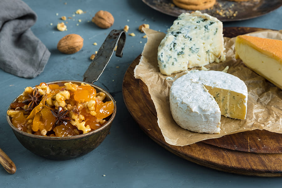 En øse med aprikoschutney med valnøtter servert på mørkt bord. Tre oster på et fat ved siden av. Hele valnøtter i bakgrunnen.