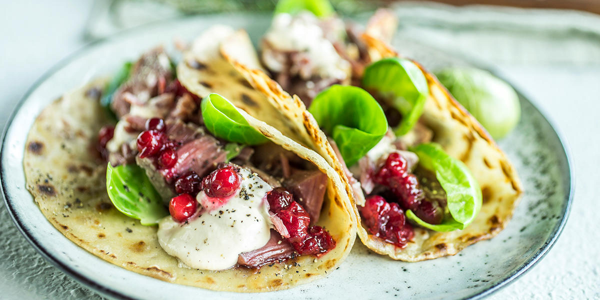 Tallerken med to lomper fylt med pinnekjøtt i biter, sennepsdressing, tyttebær og rosenkålblader.