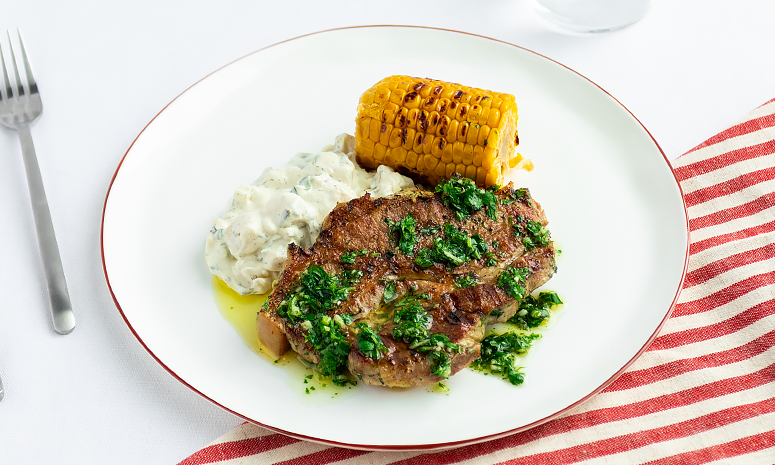 Nakkekoteletter med grillet mais, potetsalat og urtesaus servert på en hvit tallerken med et rødt og hvitt stripete håndkle under tallerkenen og en gaffel ved siden av.