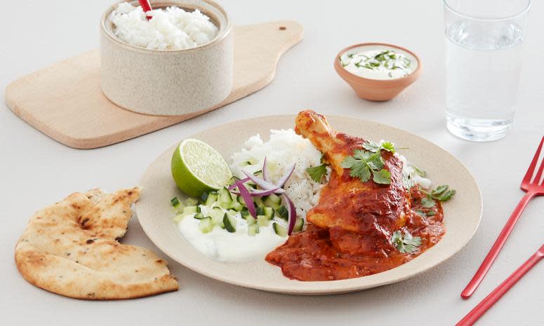Kyllinglår med tikka masala saus, ris, agurk, rødløk, limebåt og raita på en lys tallerken. Skål med ris i bakgrunnen, og naan på siden.