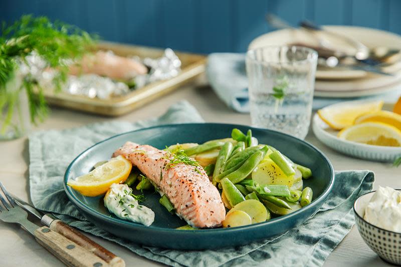 Grillet laks bakt i folie servert på mørkegrønn middagstallerken med poteter, sukkererter og brekkbønner, med sitronbåt og urtedressing på siden. Tallerkenen står på dekket middagsbord.