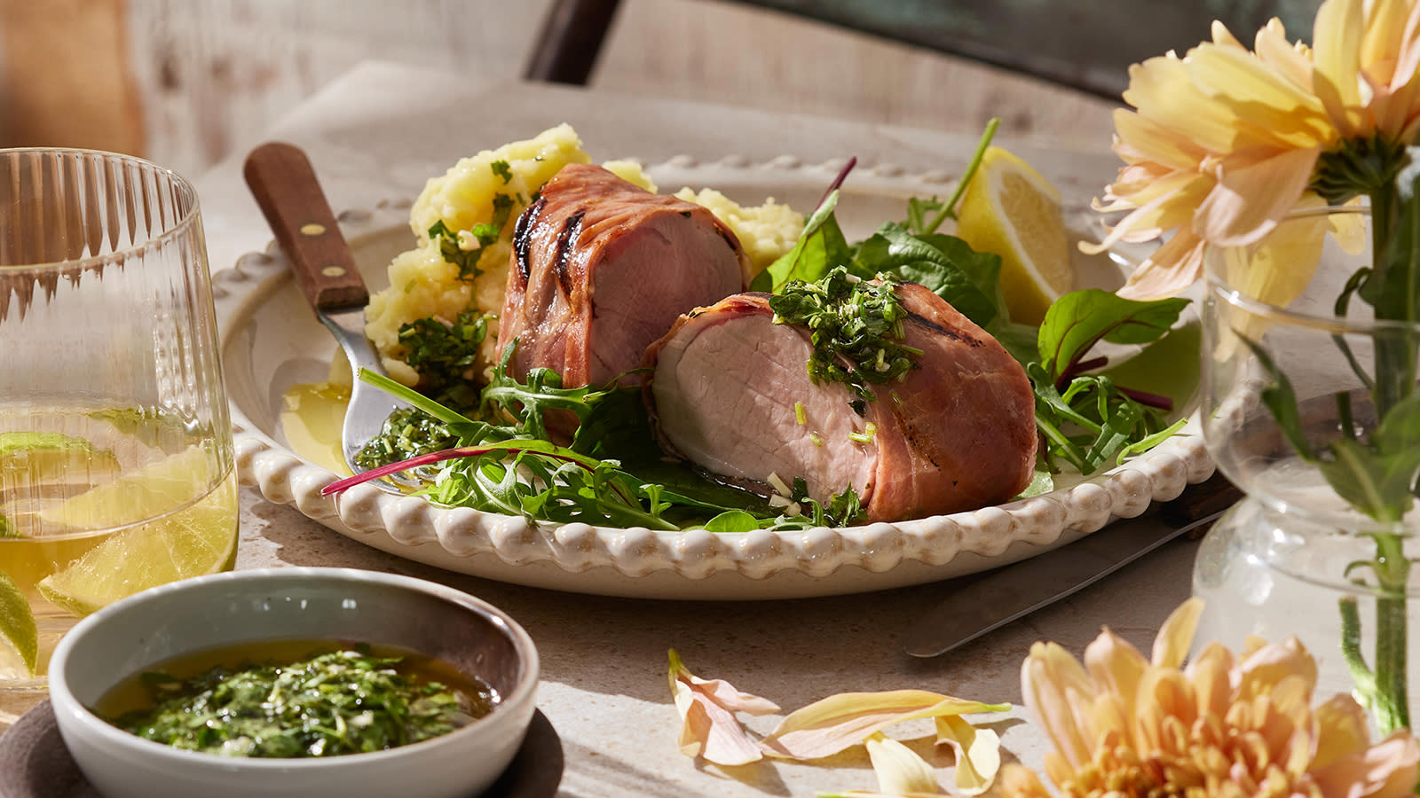  Svinefilet surret i serranoskinke servert med potetstappe og salat på dekket middagsbord med lyse blomster og et vannglass med limebåter ved siden av.