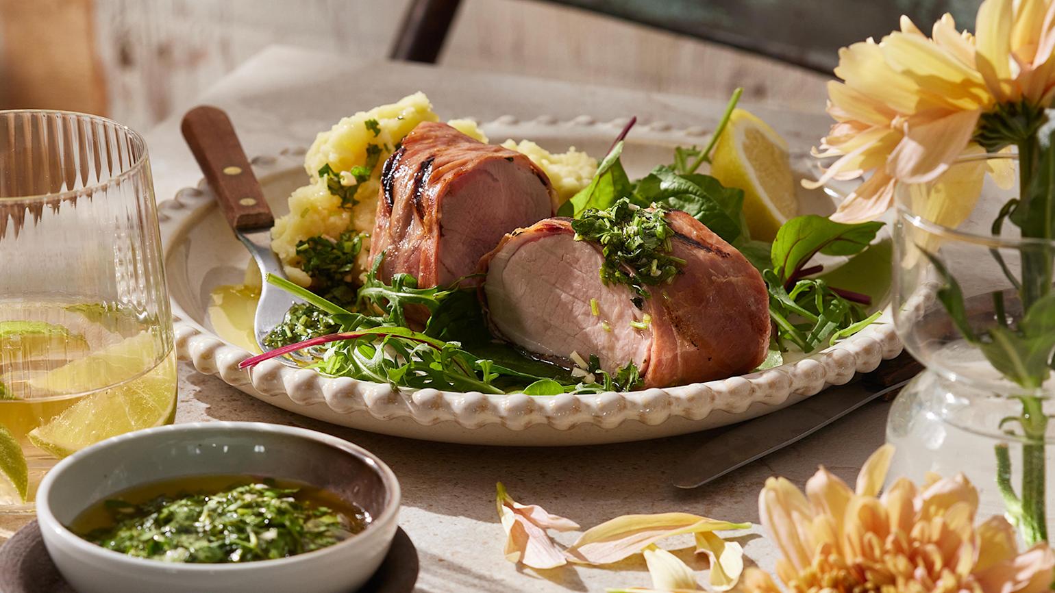  Svinefilet surret i serranoskinke servert med potetstappe og salat på dekket middagsbord med lyse blomster og et vannglass med limebåter ved siden av.