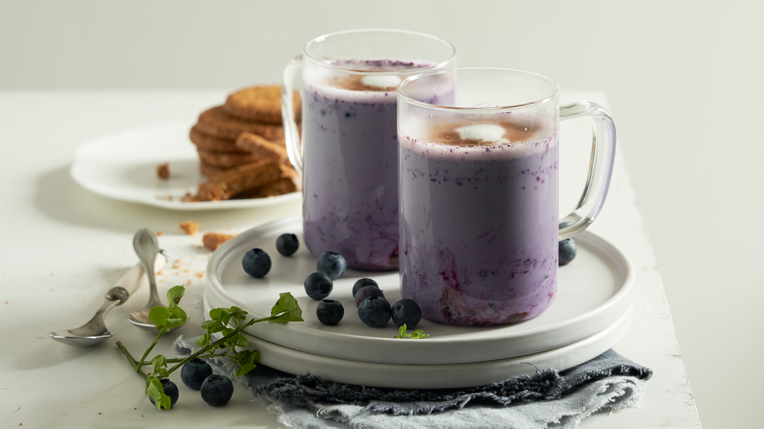 To glasskrus meg blåbærlatte servert på en tallerken med ferske blåbær rundt. Hvitt fat med havrekjeks i bakgrunnen.