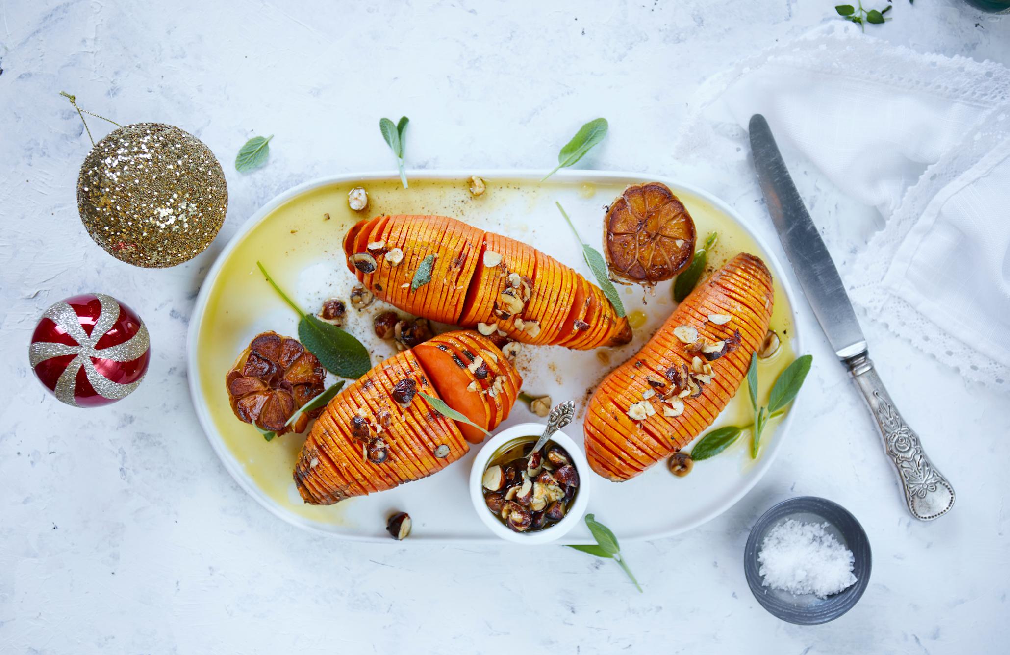 Hasselback søtpoteter på fat, med stekt hel hvitløk delt i to, overstrødd med hasselnøtter og ferske urter.