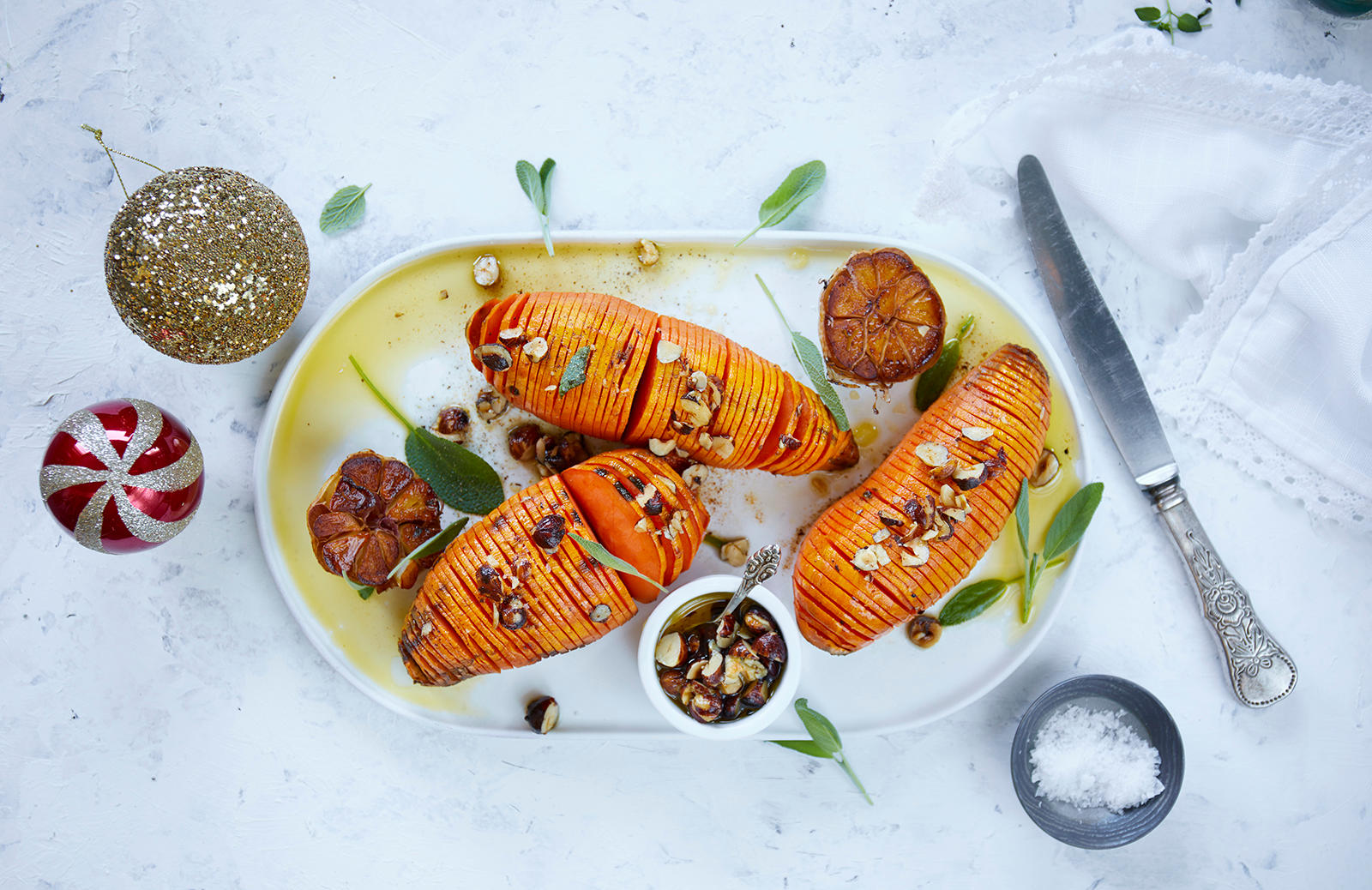 Hasselback søtpoteter på fat, med stekt hel hvitløk delt i to, overstrødd med hasselnøtter og ferske urter.