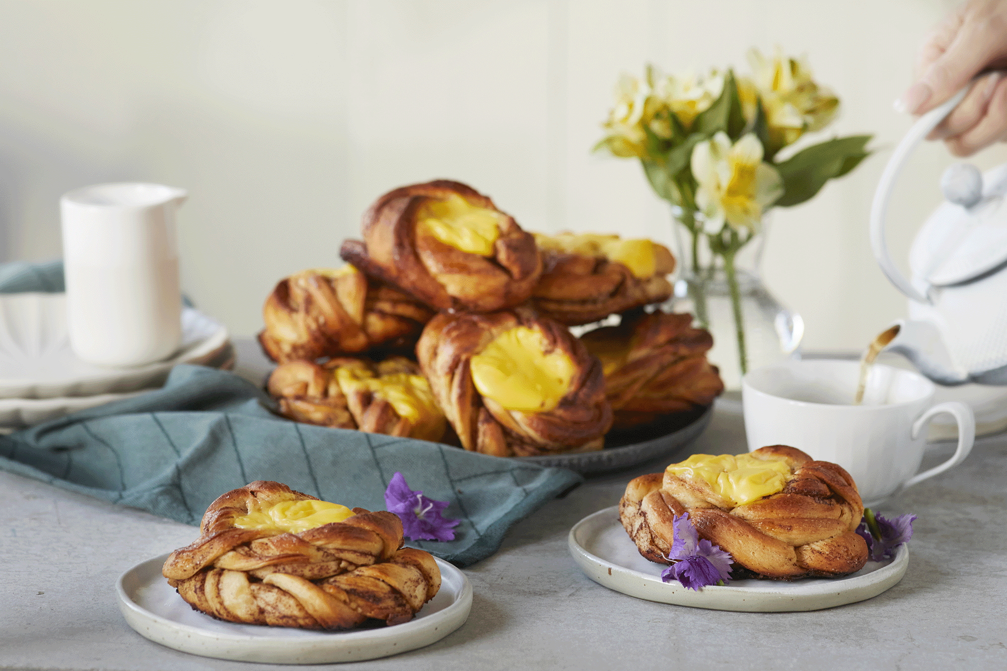 Kanelboller med vaniljekrem servert på fat og små asjetter på dekket kaffebord pyntet med fargerike blomster. Kaffe helles i en kopp ved siden av fra hvit kaffekanne.