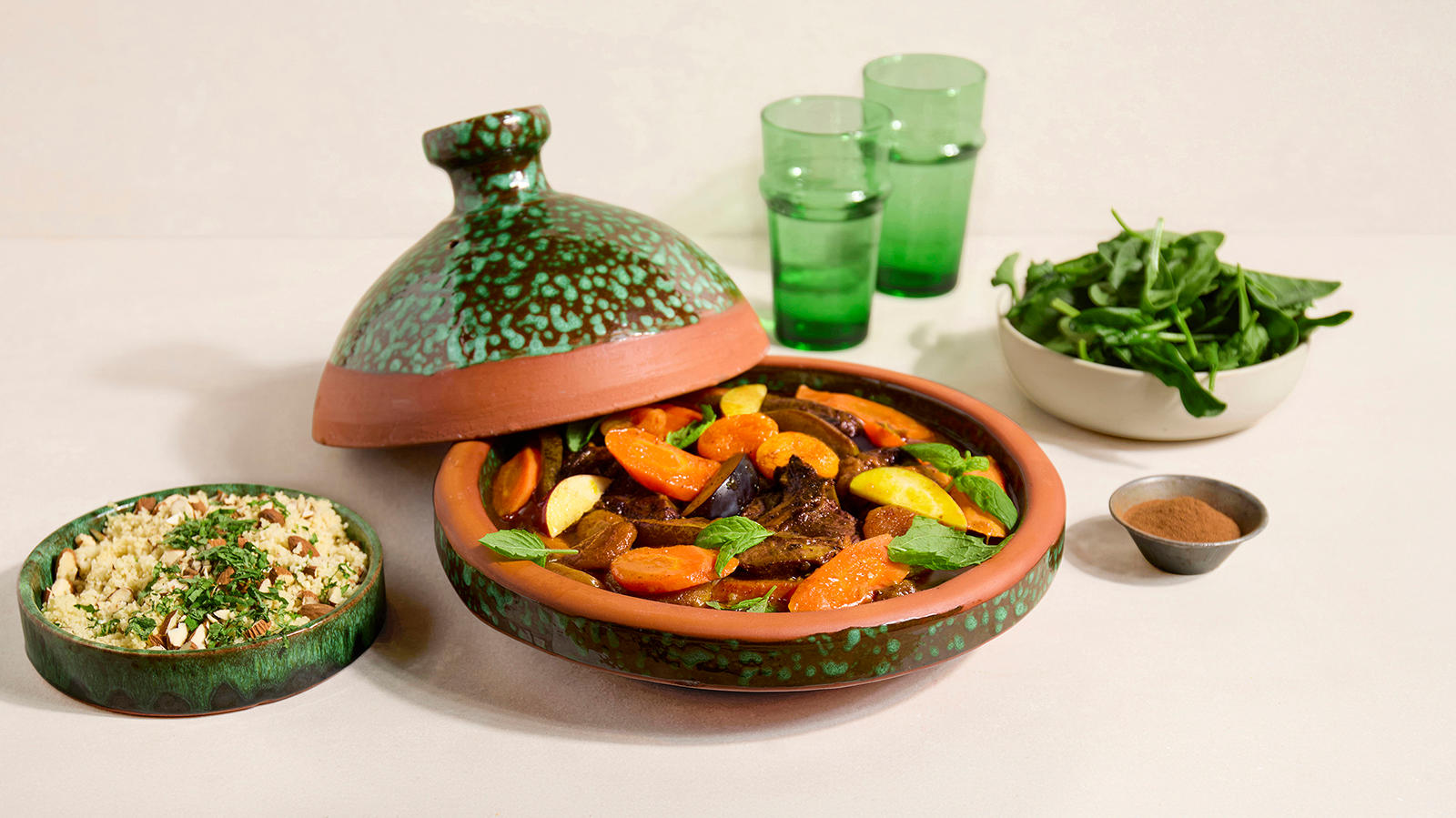 Marrokansk tagine med lam. Et fargerikt og innbydende måltid. Marrokansk lammetagine med frukt, servert i et dekorativt keramikkfat. Ved siden av står couscous med urter og nøtter, frisk salat og grønne glass som står i bakgrunnen.
