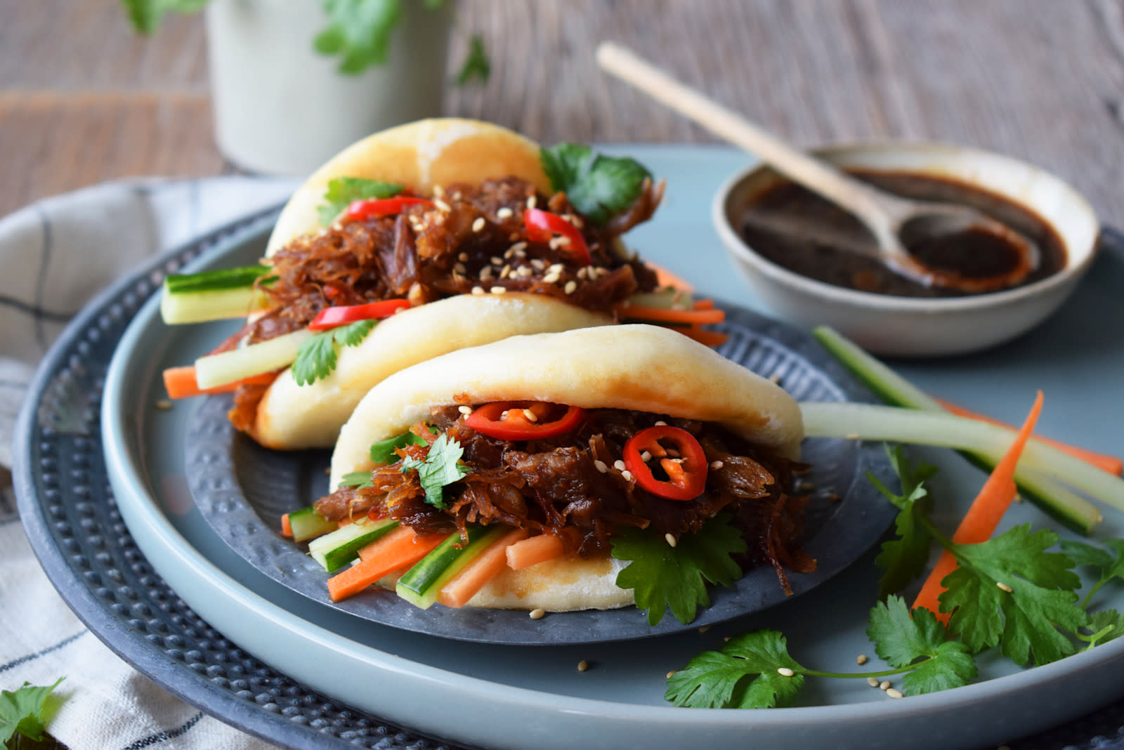 Steam buns på blått fat med soyaglasert Henriettes pinnekjøtt, strimlet agurk og gulrot, fersk koriander og chili. Sesamfrø strødd over, og ekstra soyadip på siden. Rustikk bakgrunn.
