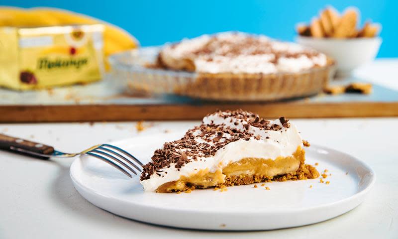 Banoffee pie, pai med kjeksbunn, karamellfyll, banan og pisket krem med revet sjokolade på toppen. En bit med pai servert på hvit tallerken.