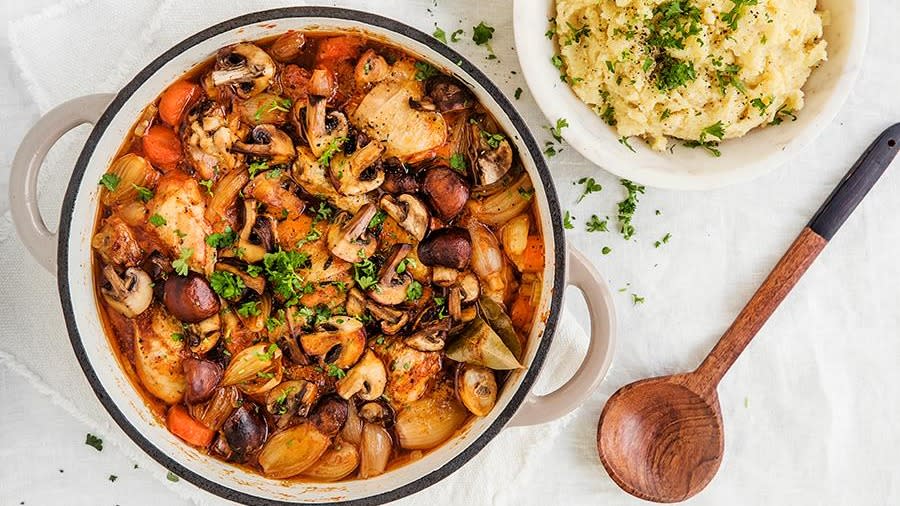 Langtidskokt kyllinggryte med sopp og bacon, småløk og gulrot servert i beige støpejernsgryte med en skål potetmos på siden.