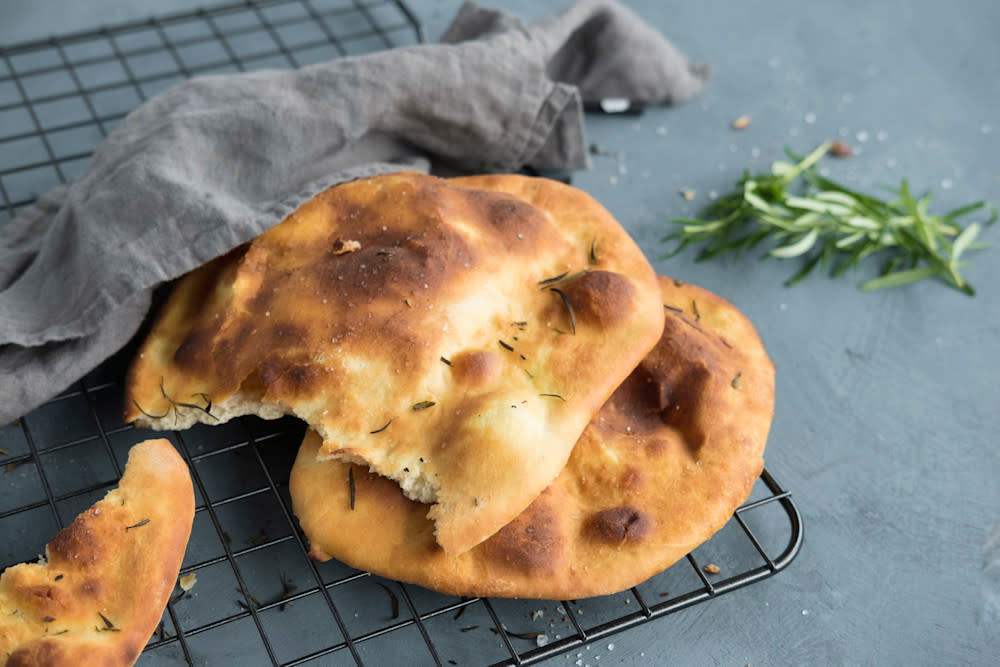 Italiensk flatbrød med rosmarinolje stablet på kjølerist med mørkegrått linhåndkle halvveis over. Frisk rosmarin ved siden av.
