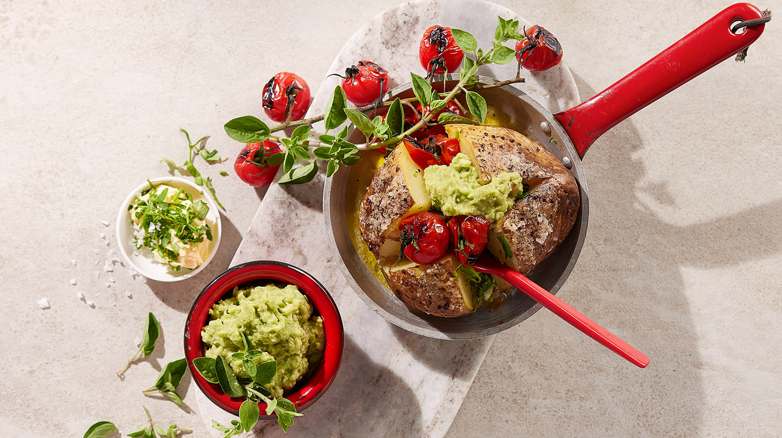 Bakt potet med urtesmør, guacamole og grillede cherrytomater servert i en kjele på lys marmorfjøl. Små skåler med guacamole, urtesmør og en ranke grillede cherrytomater på siden.