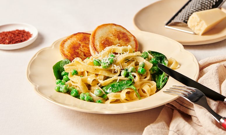 Fiskekaker med pasta og grønnsaker i fløtesaus servert i en dyp off-white tallerken med bestikk på høyre side, og parmesan på en tallerken i bakgrunnen
