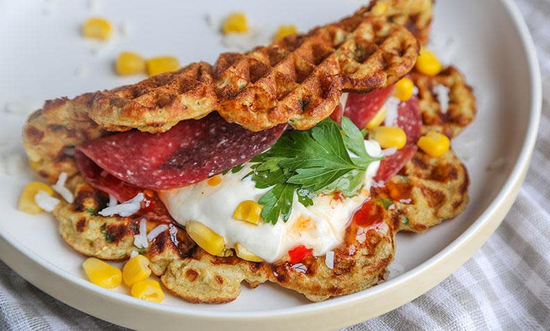 Middagsvafler med ris og mais i røren, servert med maiskorn, rømme, salami og chilisaus, på hvit tallerken.