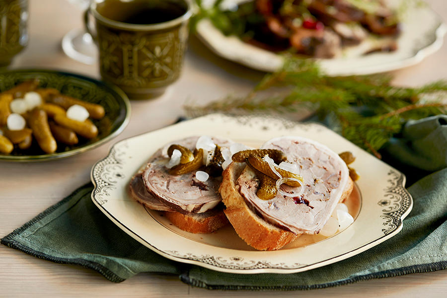 To brødskiver med hjemmelaget lammerull, toppet med syltet agurk og løk, servert på gammeldags servise. Grønn serviett, og granbar.