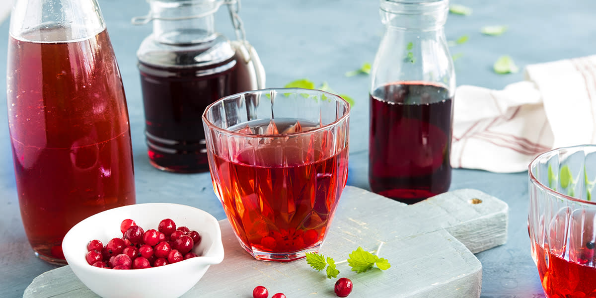 Hjemmelaget ripssaft i glass på trefjøl med liten hvit skål friske rips ved siden av. Forskjellige typer flasker med ripssaft i ulike sjatteringer i bakgrunnen.