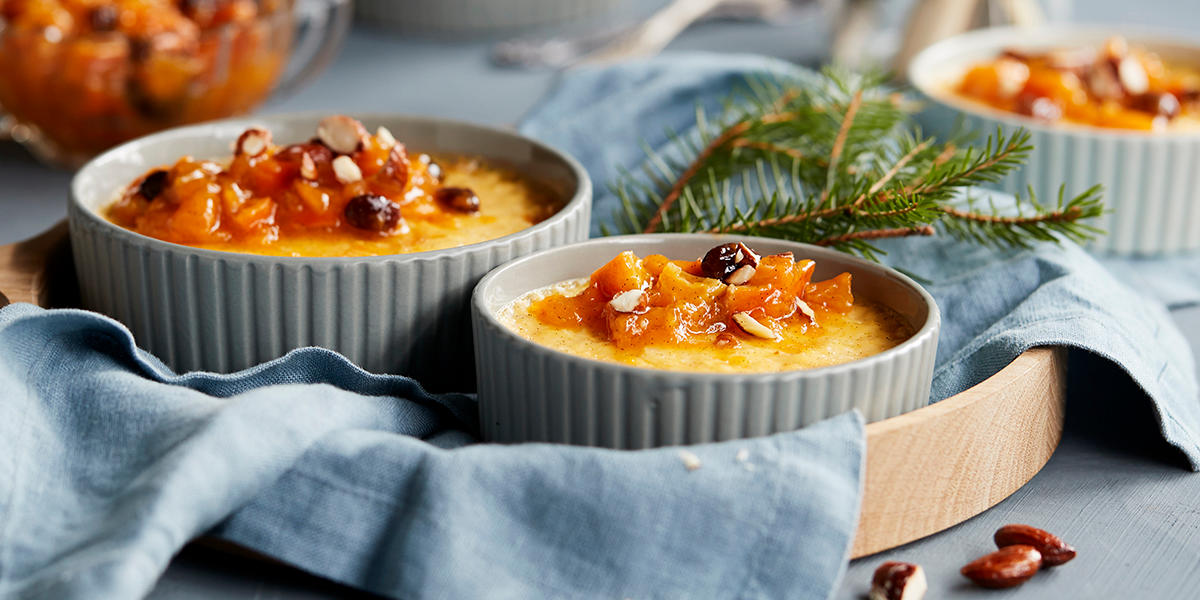 Rispudding med krydret klementinkompott og honningristede mandler servert i ildfaste grå skåler på lyst trefat dekket med lyseblå linserviett. Granbar ved siden av, og skål med krydret klementinkompott i bakgrunnen.