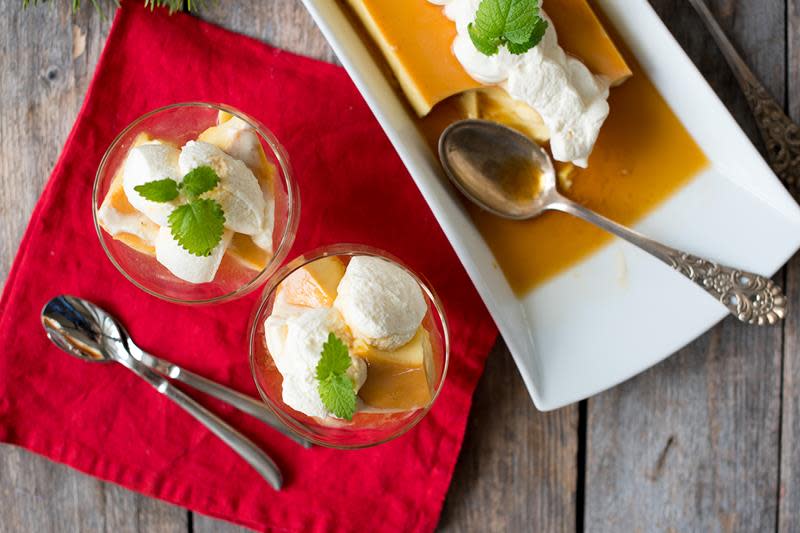 Hjemmelaget karamellpudding servert i små dessertglass, toppet med pisket krem og sitronmelisse. Rød linserviett på rustikk trebakgrunn. Serveringsfat med hel karamellpudding ved siden av.