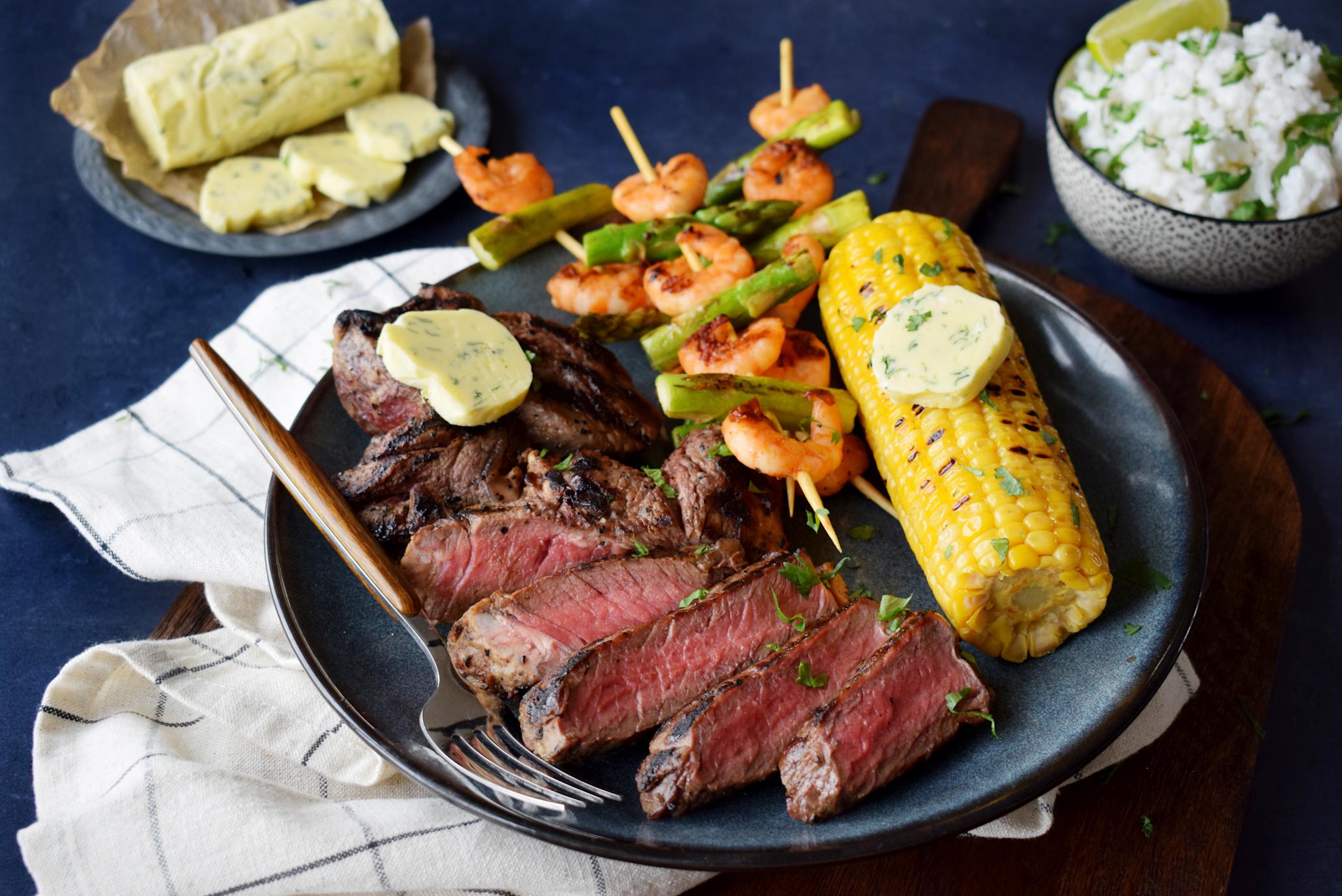Surf'n turf entrecôte med scampispyd med asparges, og grillet maiskolbe med hjemmelaget lime- og koriandersmør servert på mørkeblå tallerken. Skål med kokosris, og lite fat med hjemmelaget hvitløkssmør i bakgrunnen.