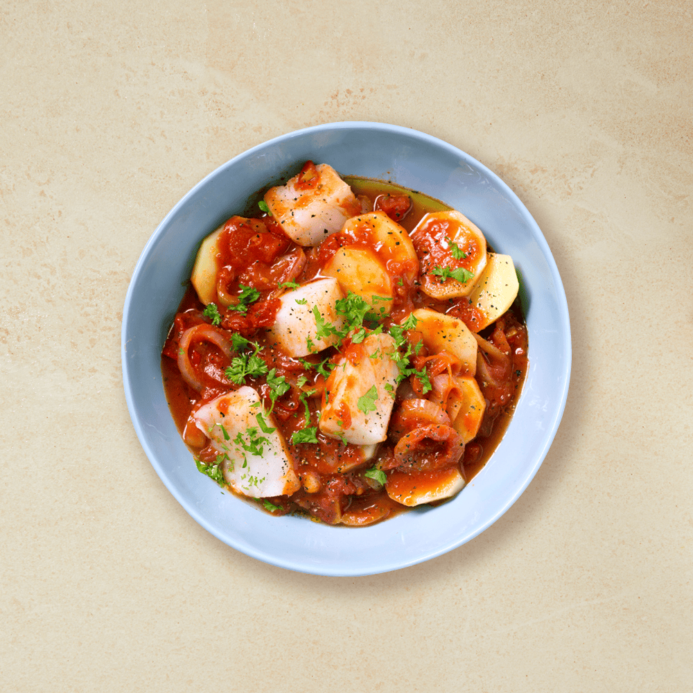 Enkel bacalao med poteter i tomatsaus pyntet med kruspersille, servert i lyseblå skål på beige bakgrunn.