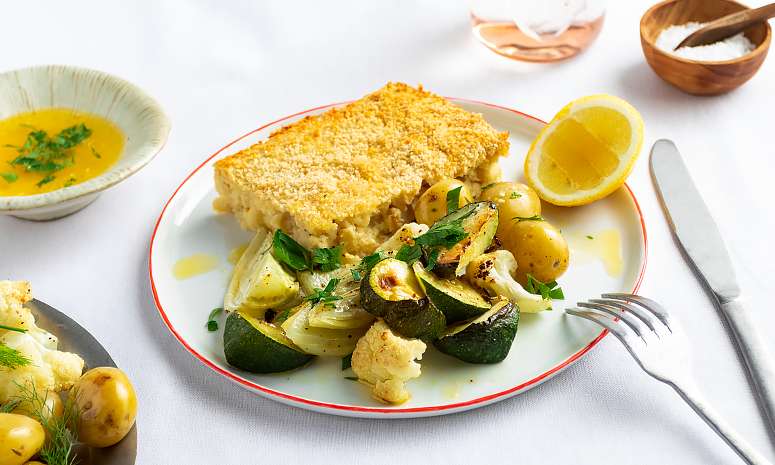 Fiskegrateng med ovnsbakte grønnsaker og sitron servert på en hvit tallerken med rød kant og bestikk på høyre side. På venstre side ligger en skål med smeltet smør 